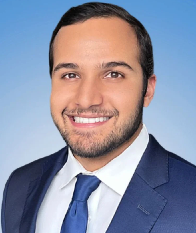 Original smiling headshot with short side-parted hair, a short beard, navy suit, white shirt and blue tie.