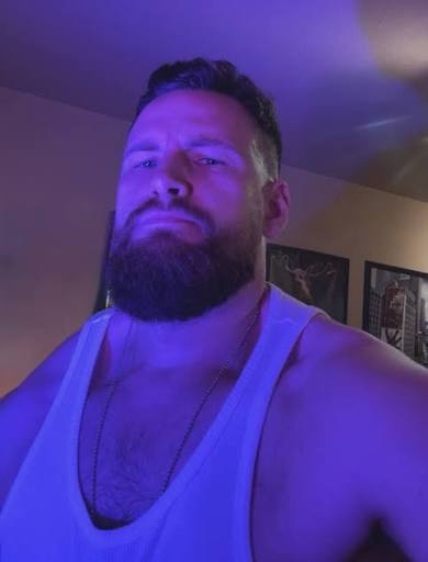 Original purple-lit selfie showing a man with a full beard, short upward-brushed hair, pale sleeveless top and chain.
