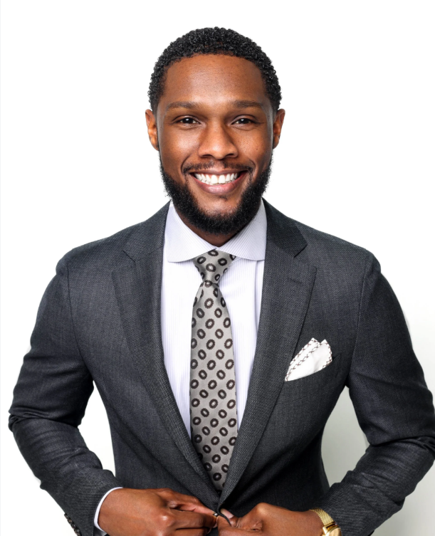 Original smiling waist-up portrait with short natural curls, a beard, charcoal suit, patterned tie and hands near the jacket fastening.